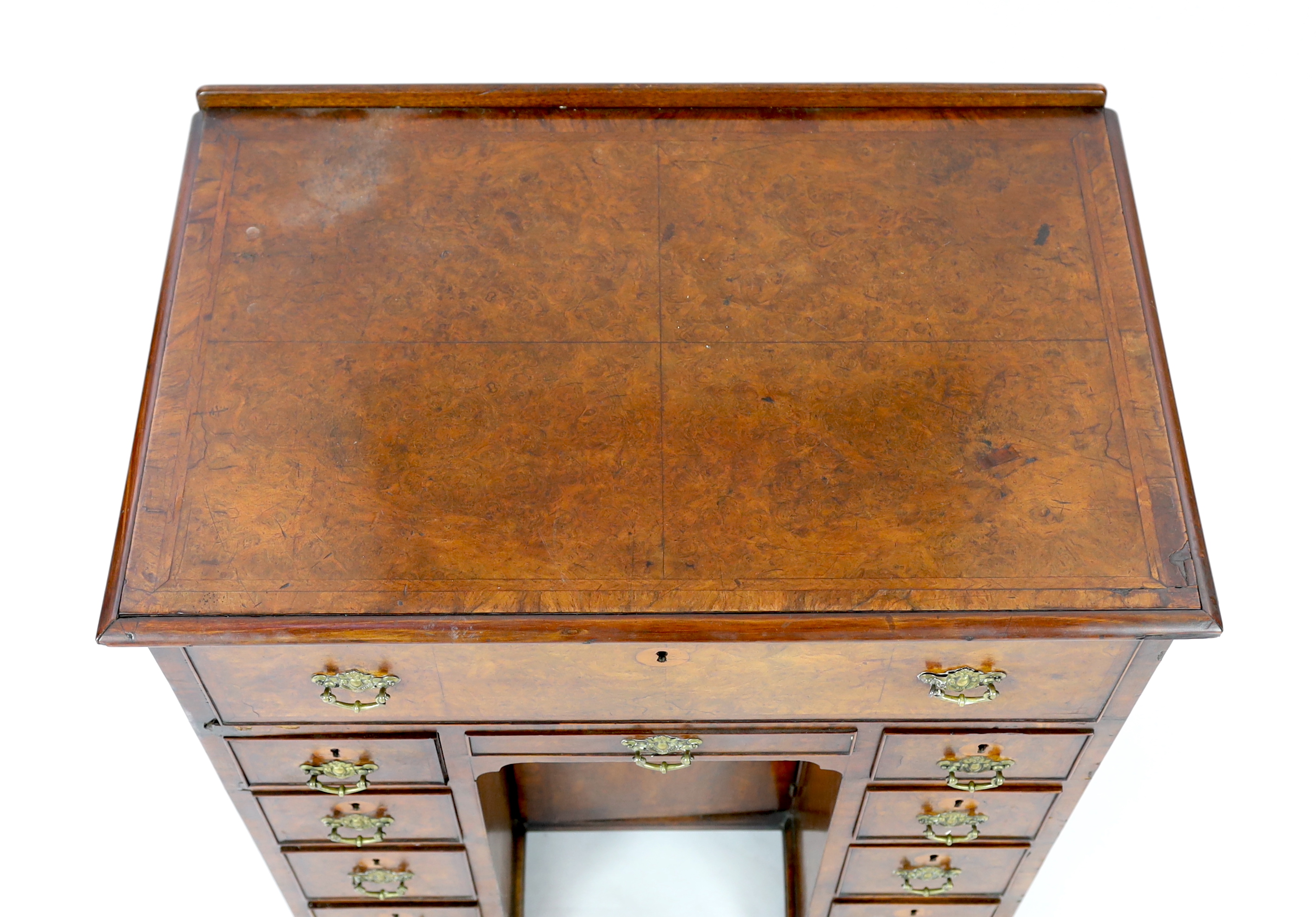 An early 18th century walnut kneehole secretaire desk, with quarter veneered top and fall front drawer opening to reveal pigeon holes and drawers, over eight further drawers surrounding a central recessed cupboard, on pa